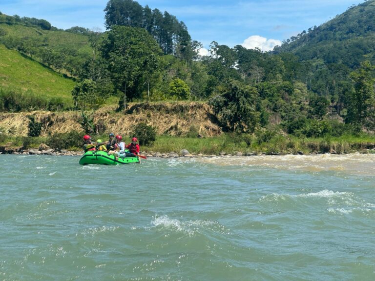A green raft with five paddlers in red helmets floats down the wide, sunny Pejivalle River between green farm hills