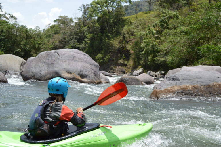 A kayaker in a blue helmet and lime-green kayak paddles toward huge boulders on a sunny jungle river