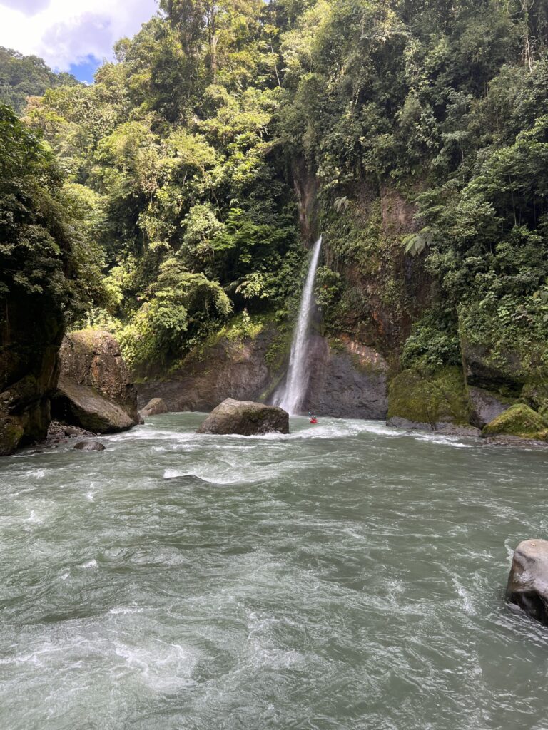 A tall waterfall pours into a wide, churning jungle river, a tiny red kayak near its base