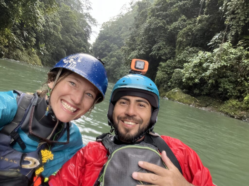 Kayla and Manuel, smiling in whitewater helmets and paddling jackets, on a green river in a jungle canyon