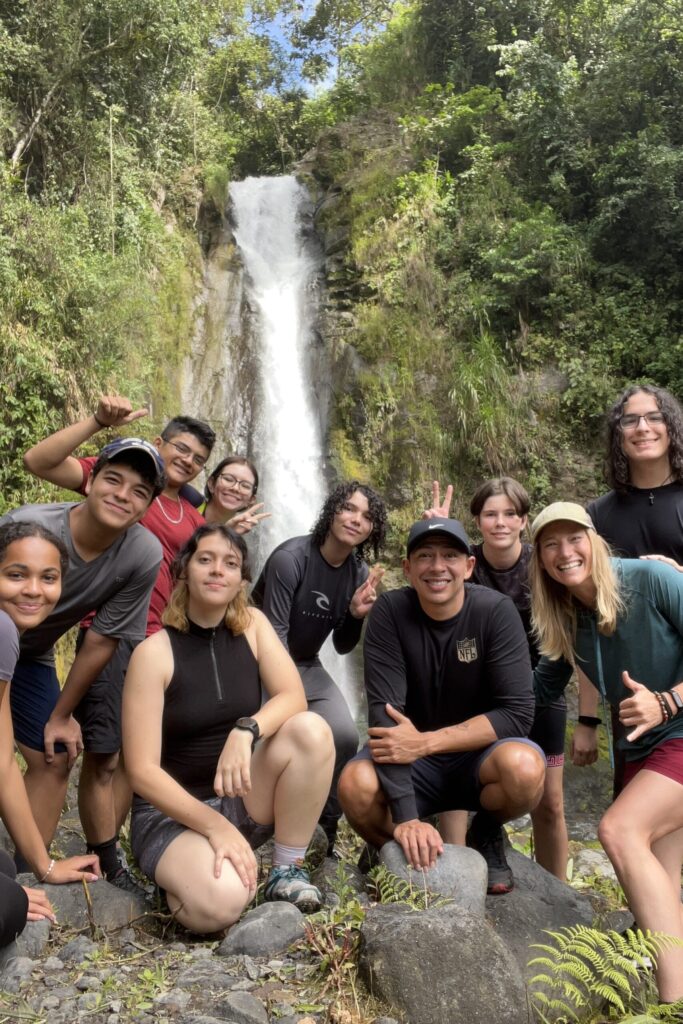 About a dozen smiling teens and young adults pose on rocks at the foot of a tall jungle waterfall