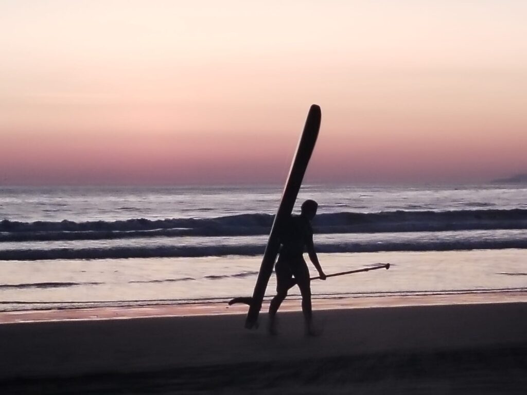 Silhouette of a person carrying a stand-up paddleboard and paddle along a beach at dusk, waves behind