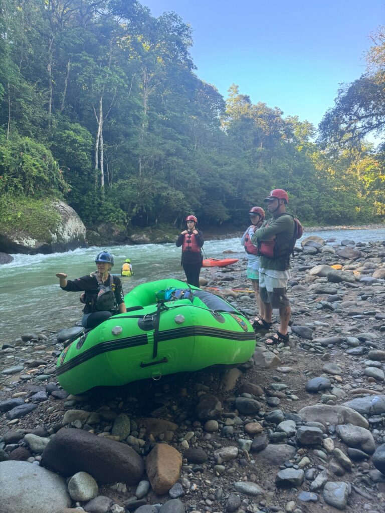 A guide in a blue helmet gives a safety talk beside a green raft as three guests in red helmets listen by the Pejivalle River
