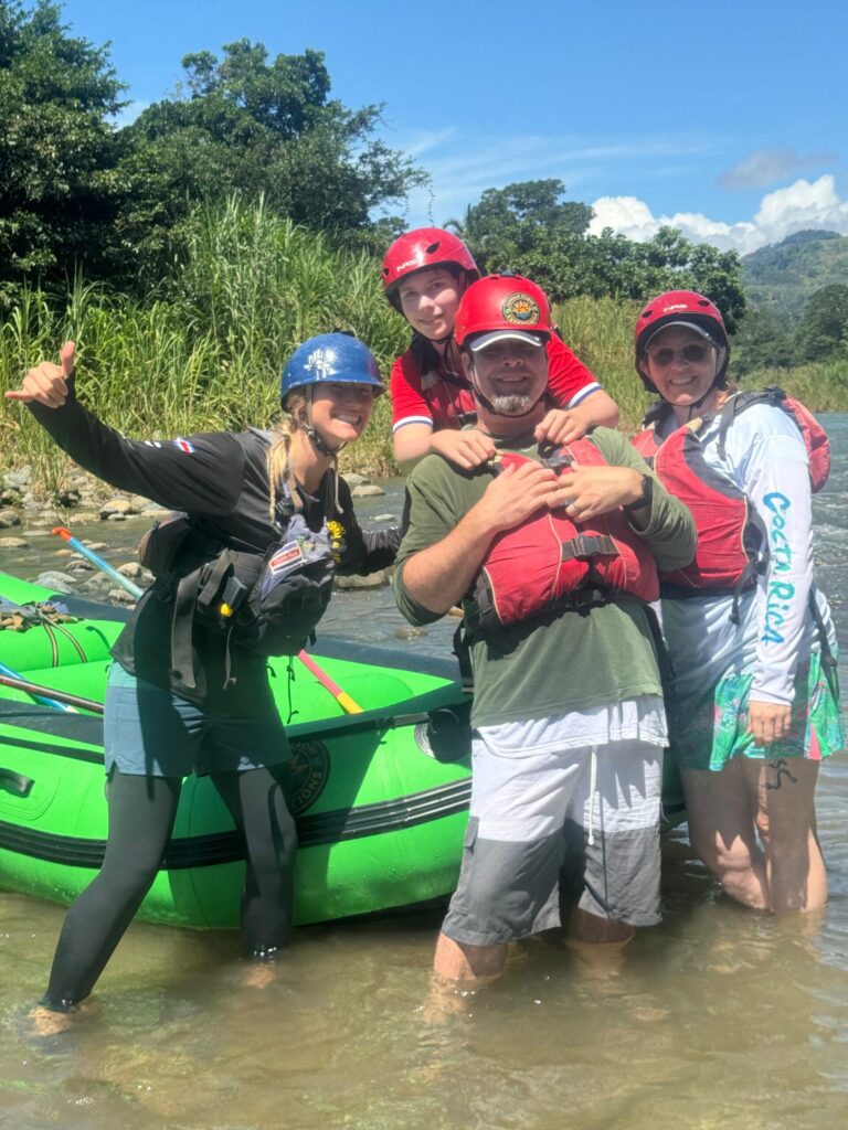 A guide and a smiling family in red helmets pose in the shallows of the Pejivalle River beside a green raft