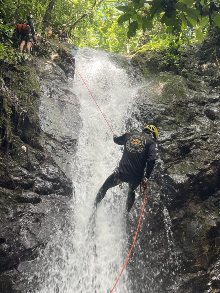 A canyoneer in a yellow helmet rappels down a rushing waterfall on an orange rope as two people wait at the top
