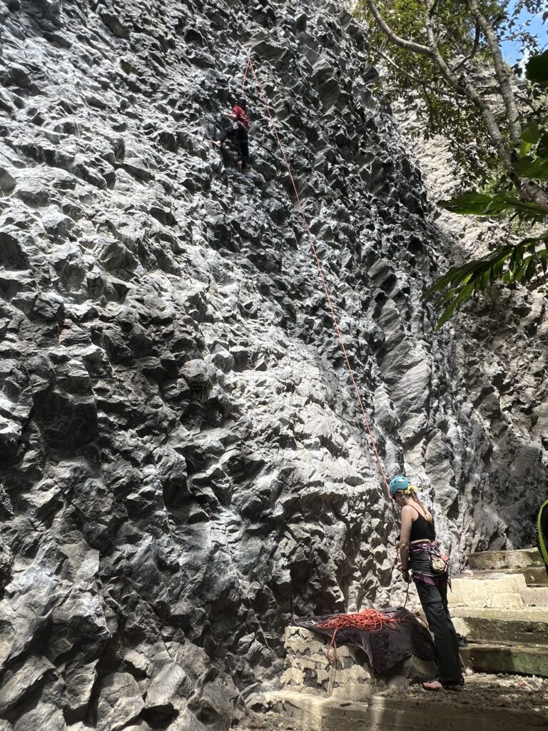 A belayer in a teal helmet feeds rope to a climber high on the towering grey rock wall at Cachí
