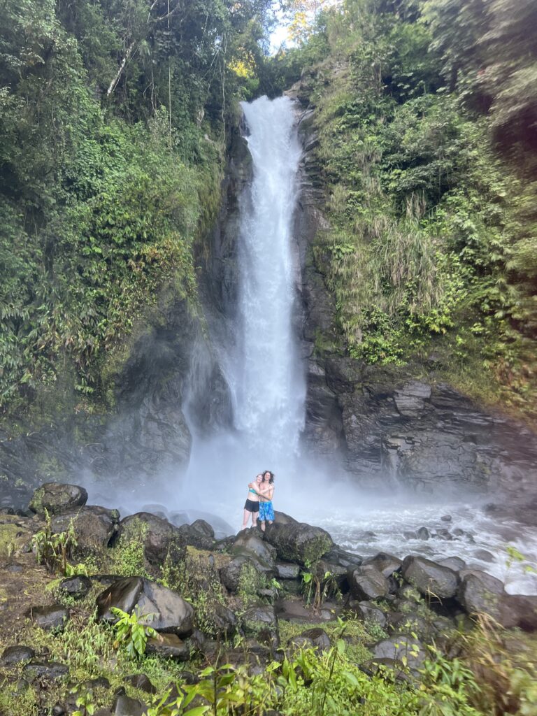 Two people stand arm in arm on boulders at the misty foot of a tall, powerful jungle waterfall