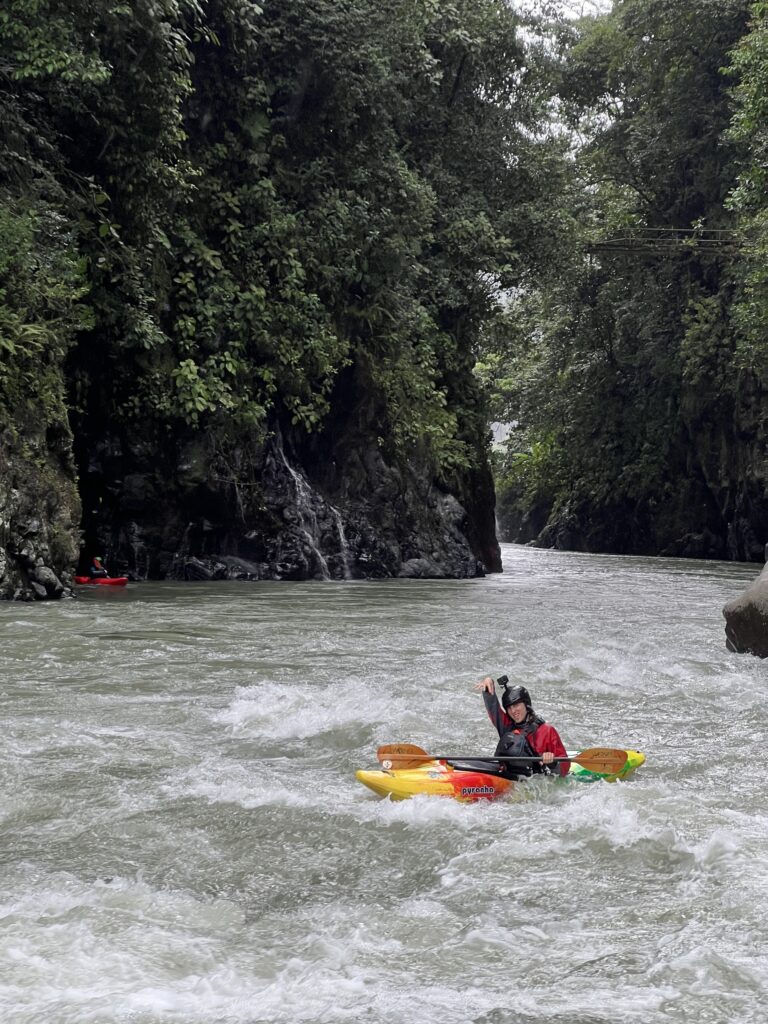 A kayaker in a red and yellow boat raises a paddle in rough water at the mouth of a steep jungle gorge