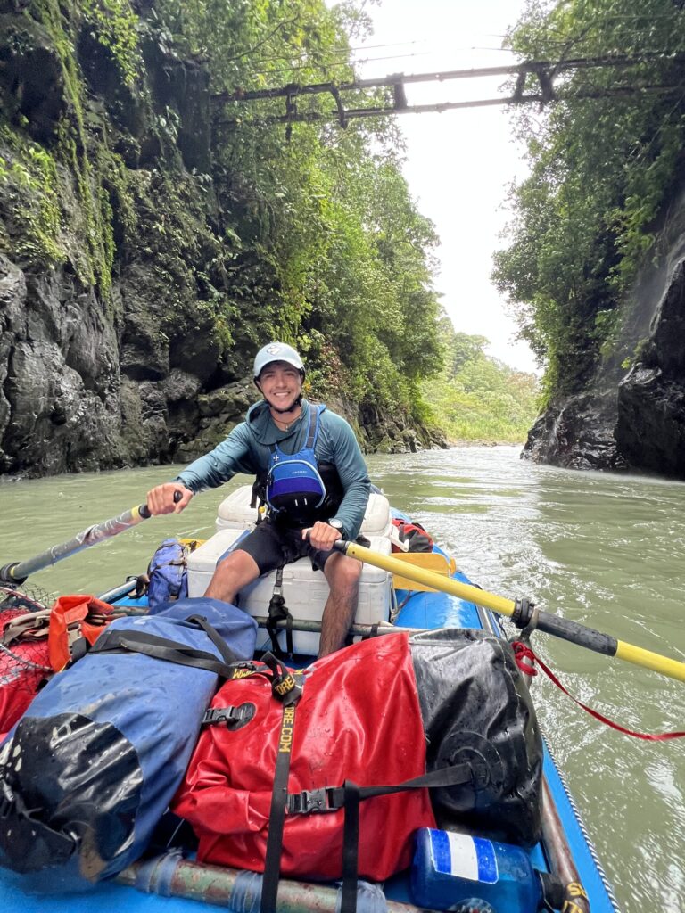 A smiling guide rows a gear raft piled with dry bags through a jungle gorge on the Pacuare River, a footbridge overhead