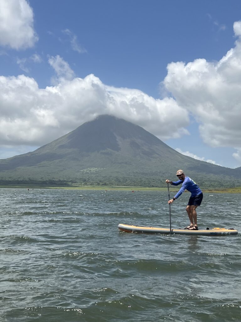 A stand-up paddleboarder on a wind-rippled lake below a cone-shaped volcano capped with cloud
