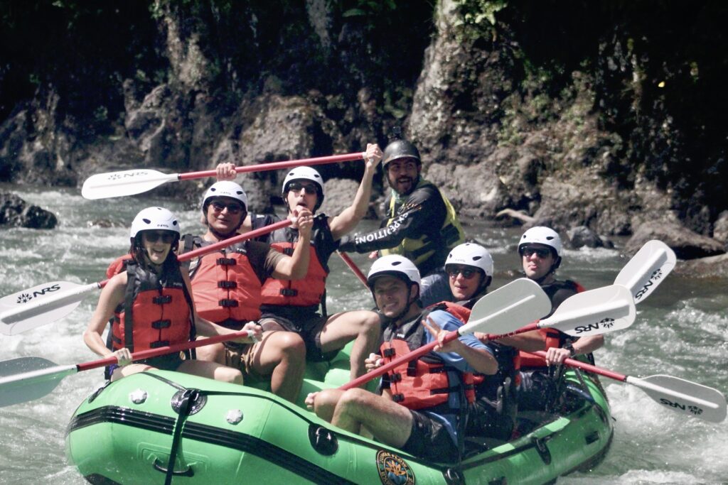 Six smiling rafters in white helmets raise their paddles in a green raft on the Pacuare River, their guide behind