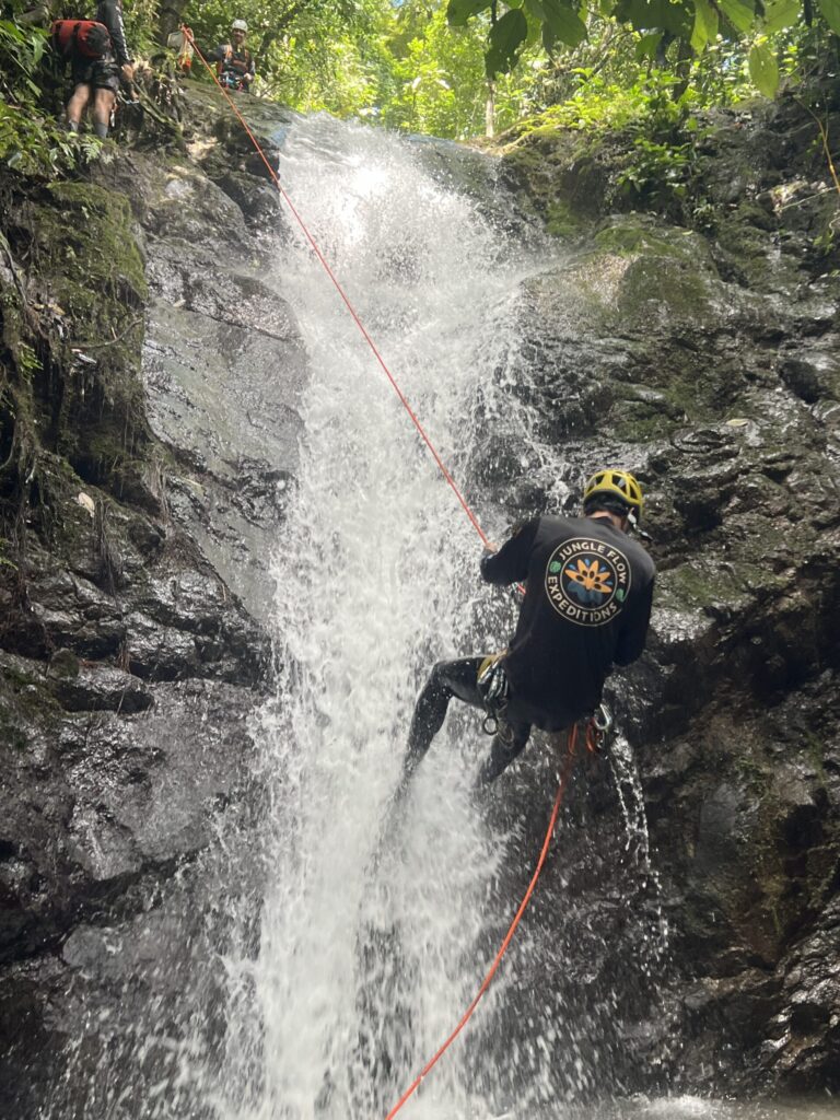 A canyoneer in a yellow helmet and Jungle Flow shirt rappels down a rushing waterfall on an orange rope