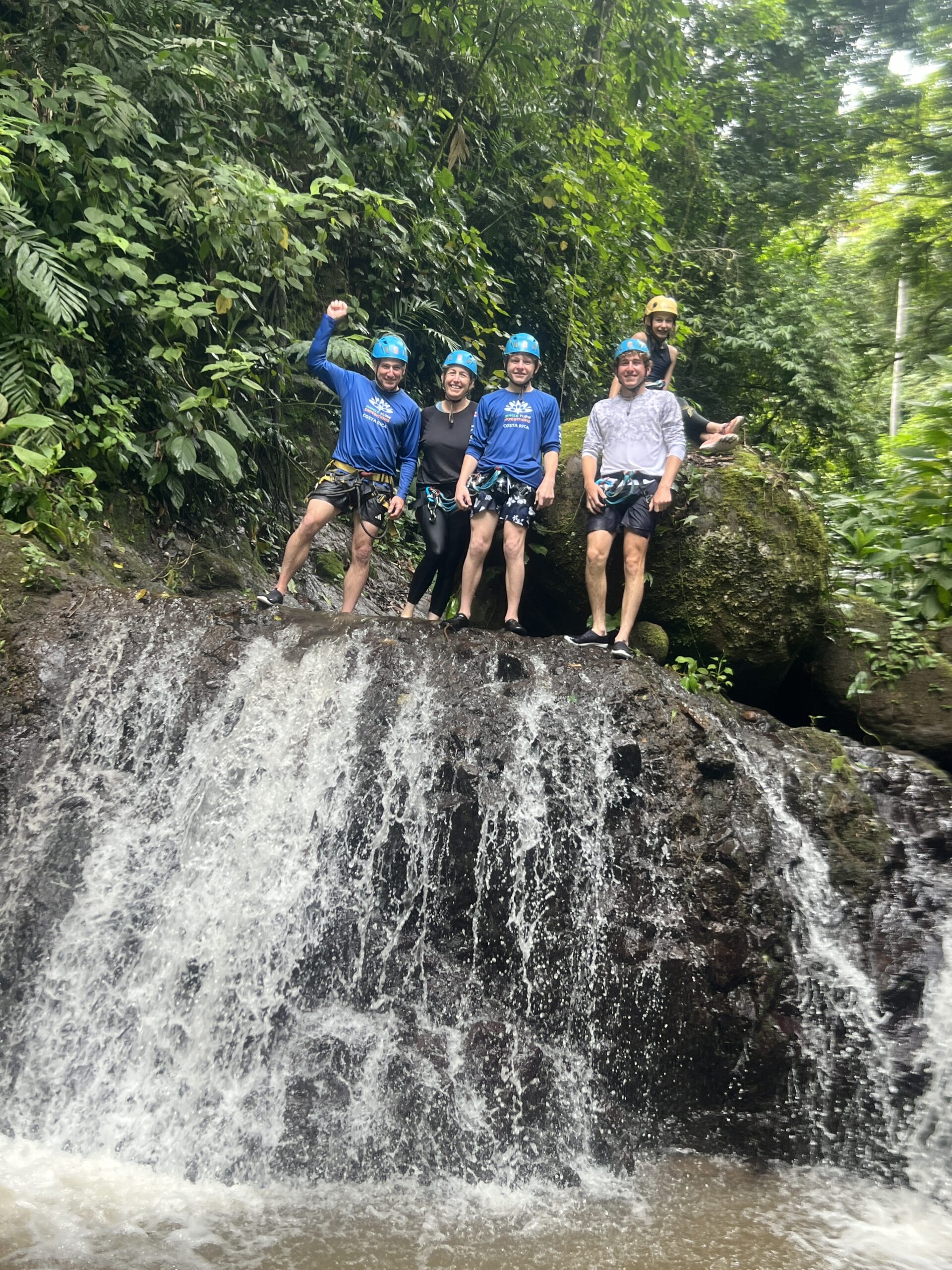 Five canyoneers in blue helmets and harnesses pose on a rock ledge above a wide jungle waterfall