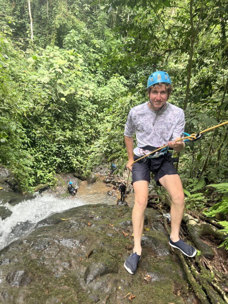 A smiling young man in a blue helmet leans back on his rope at the top of a small jungle waterfall