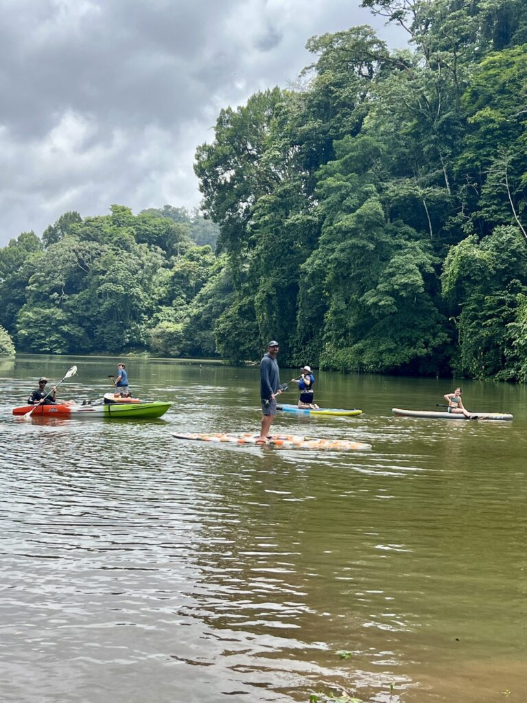 Paddleboarders and a kayaker spread out across calm Laguna Yaku Wasi, ringed by rainforest under a cloudy sky