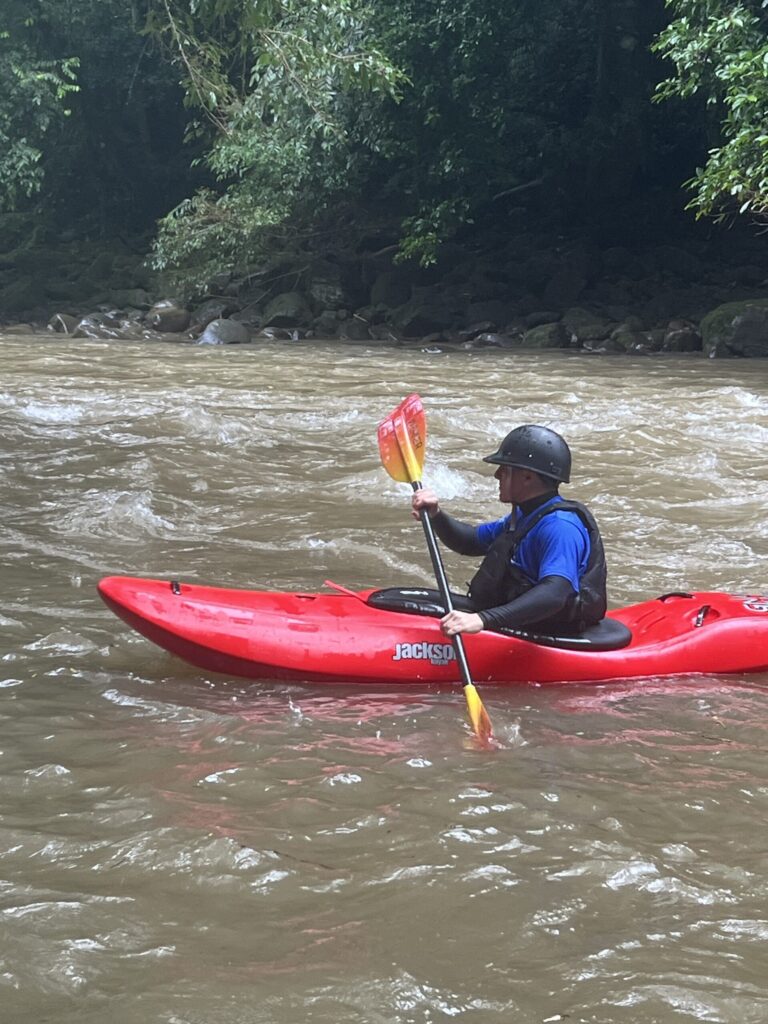 A kayaker in a black helmet paddles a red kayak across a fast brown river below a jungle bank