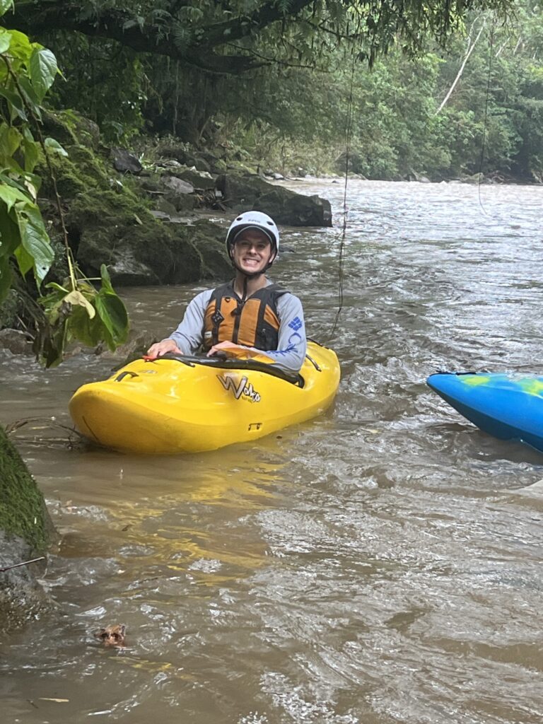 A smiling paddler in a white helmet sits in a yellow kayak in a calm eddy of a brown jungle river