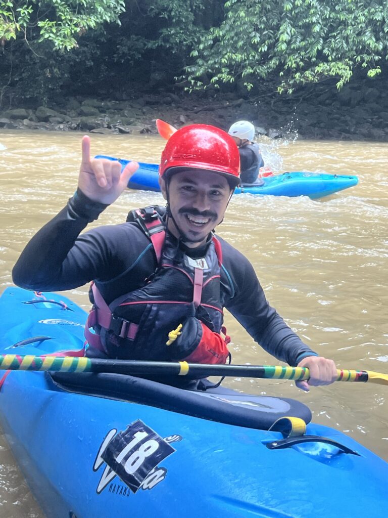 A grinning kayaker in a red helmet and blue boat makes a hand sign on a brown jungle river, another paddler behind