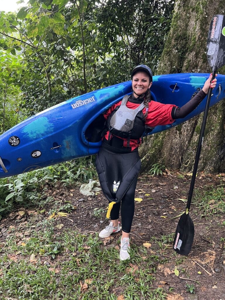 A smiling woman in a red paddling jacket shoulders a blue kayak and raises her paddle on a forest trail