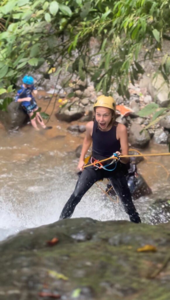 A delighted girl in a yellow helmet leans back on her rope at the lip of a waterfall while a child watches