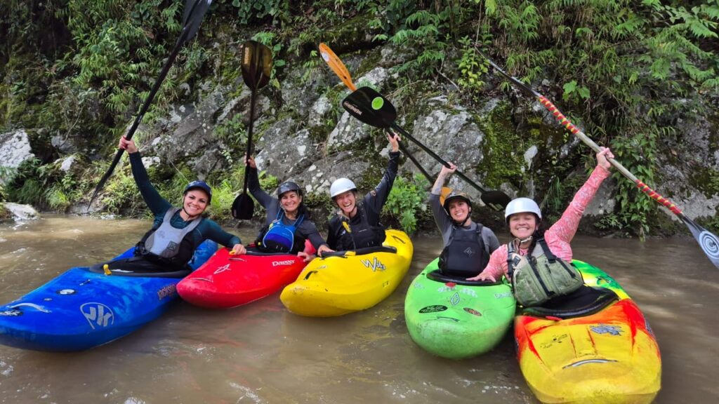 Five smiling women raise their paddles from a row of brightly colored kayaks on a brown jungle river
