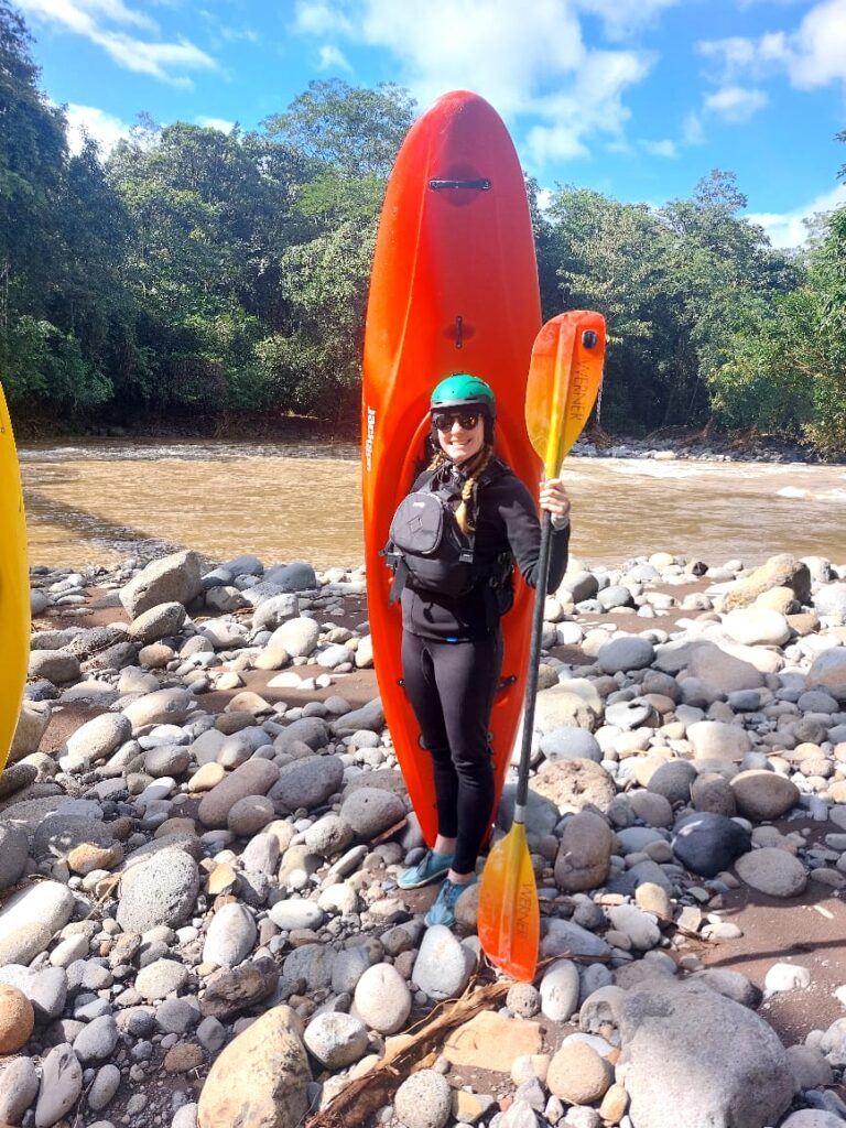 A smiling kayaker in a green helmet and sunglasses stands on a rocky riverbank before her upended orange kayak