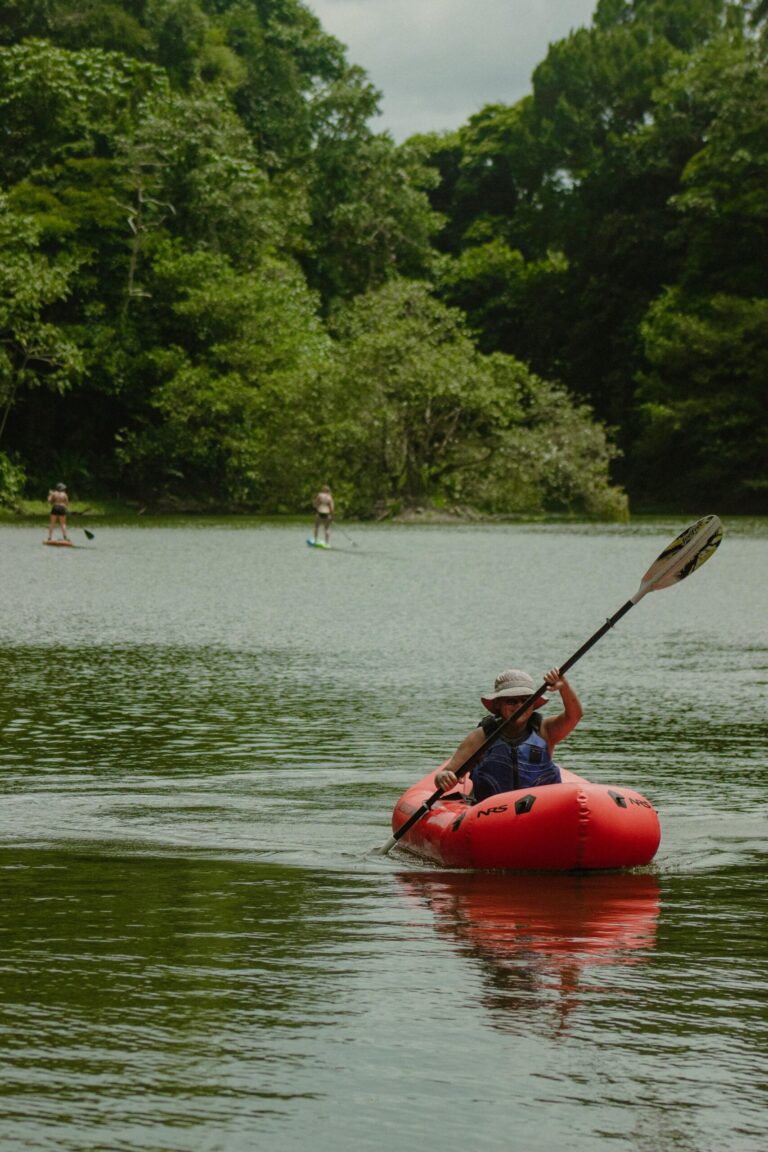 A paddler in a sun hat steers a red inflatable kayak across calm Laguna Yaku Wasi, two paddleboarders behind