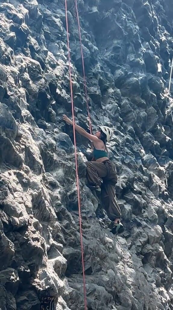A climber in a white helmet reaches for a hold on the dark, sculpted rock face at Cachí, a red rope beside her