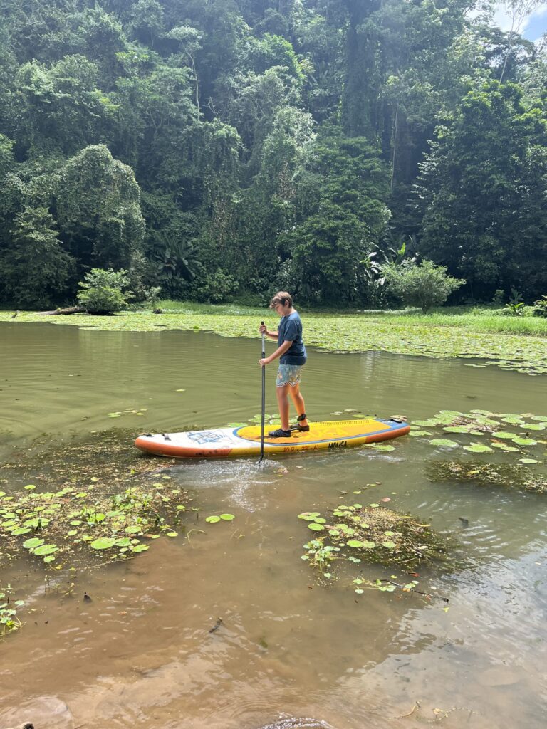 A boy on a yellow paddleboard pushes off through lily pads at the edge of Laguna Yaku Wasi, rainforest behind