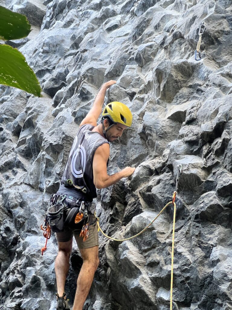 A climber in a yellow helmet and a harness racked with gear reaches up the sculpted grey rock at Cachí