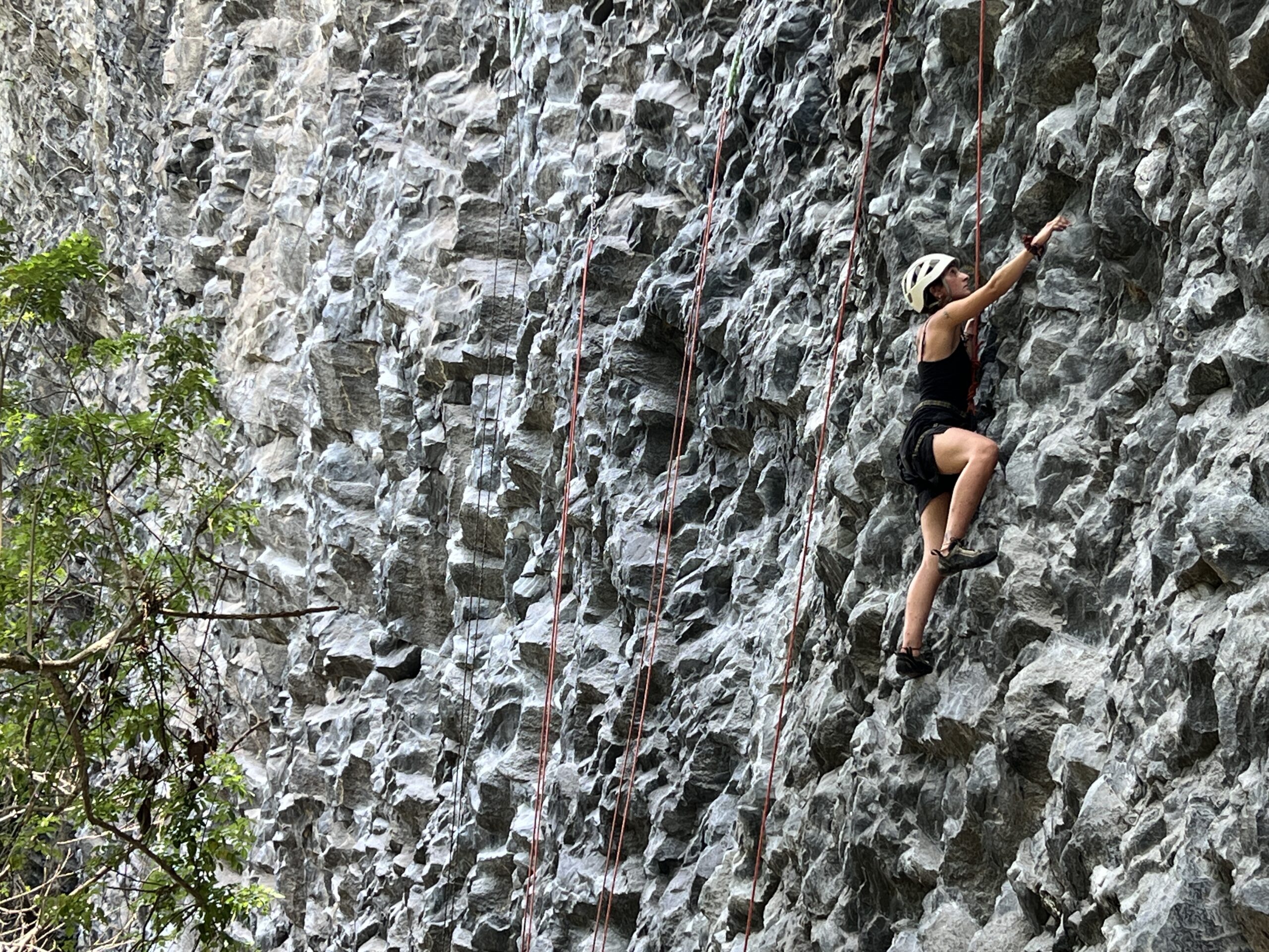 A climber in a white helmet works up the vast, sculpted grey rock wall at Cachí, red ropes hanging beside her