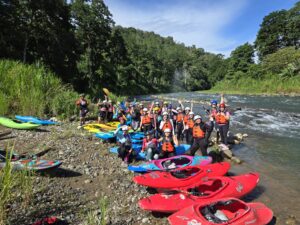 A big group of paddlers in helmets cheers beside a long row of colorful kayaks on a sunny jungle riverbank
