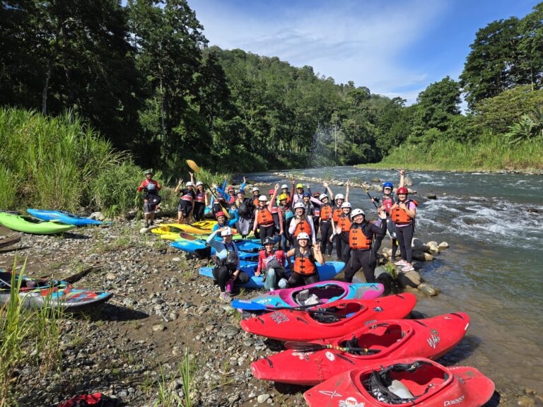 A big group of paddlers in helmets cheers beside a long row of colorful kayaks on a sunny jungle riverbank