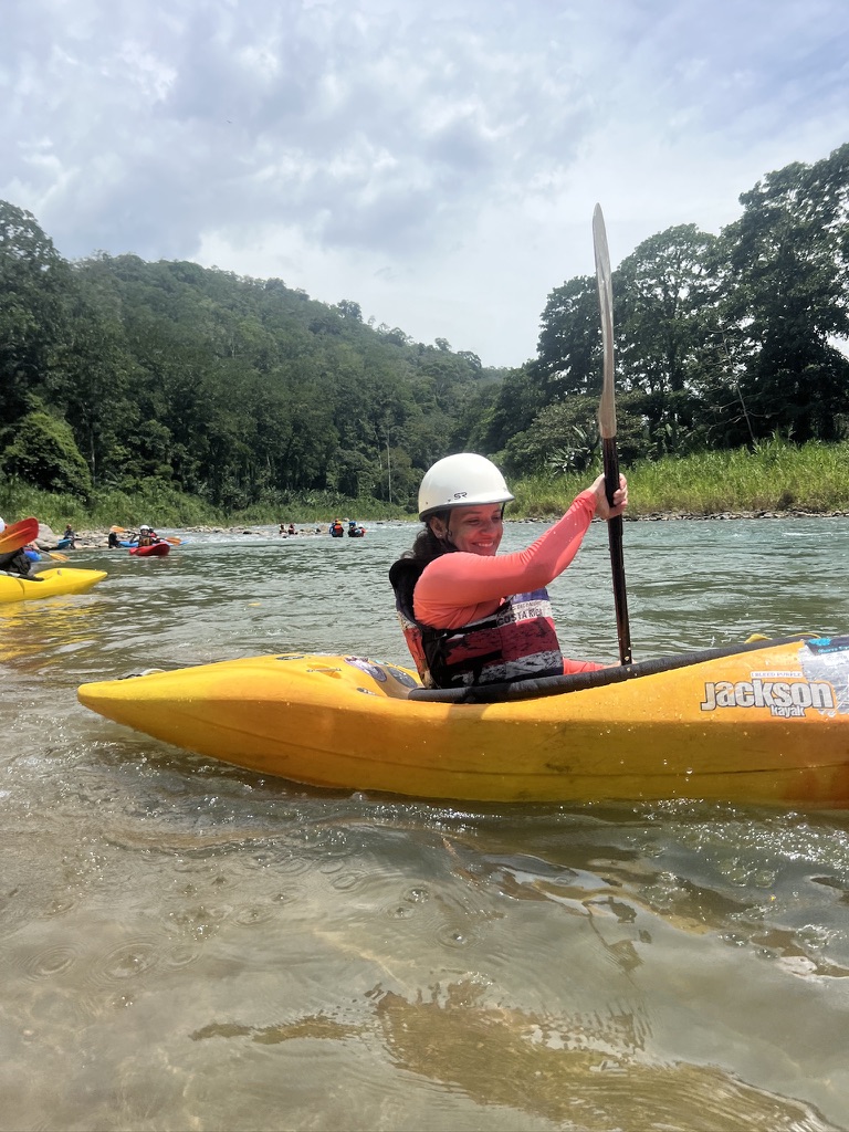 A smiling woman in a white helmet paddles a yellow kayak on a clear river, a group of kayakers behind her