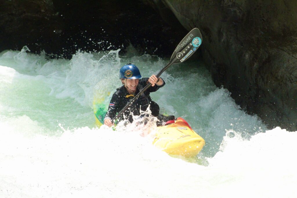 A grinning kayaker in a blue helmet drives a yellow kayak through foaming whitewater beneath a dark rock overhang