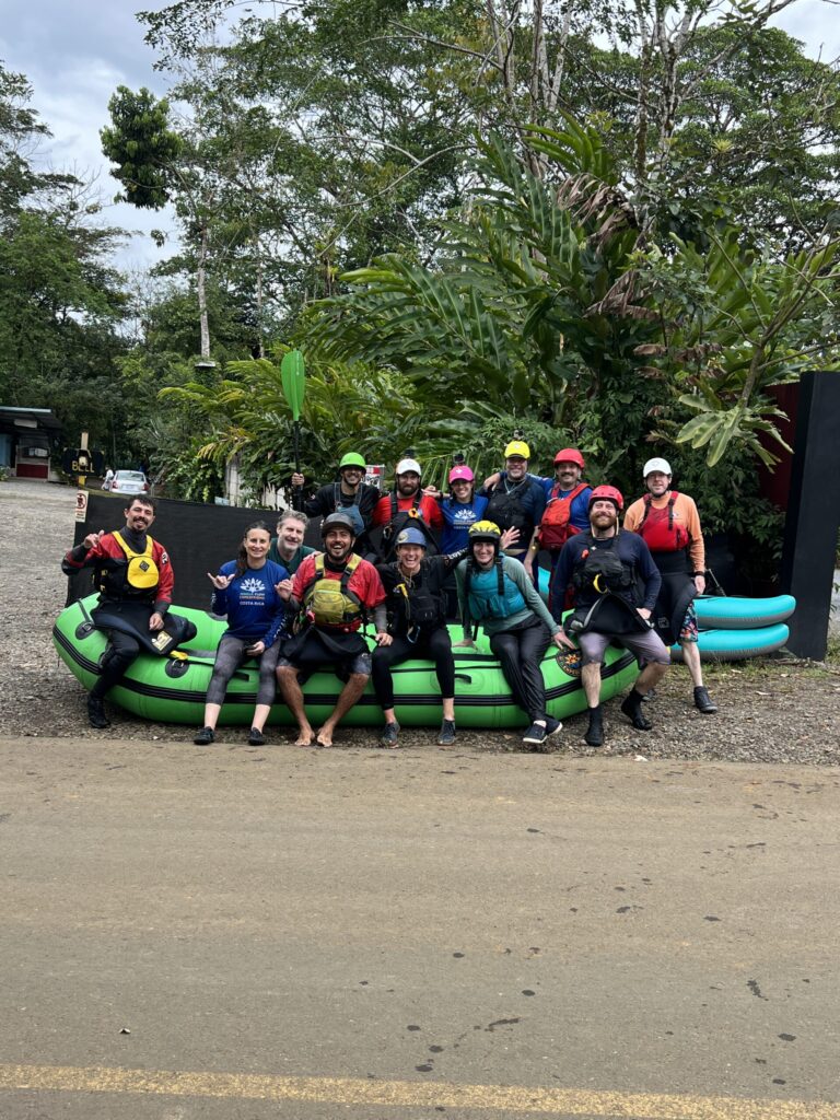 About fifteen paddlers in helmets and spray skirts pose on and around a green raft parked by a road