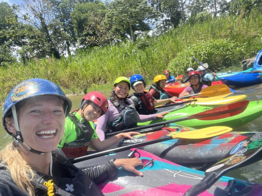 Six smiling women in helmets sit in a row of colorful kayaks on a jungle river, the nearest taking a selfie