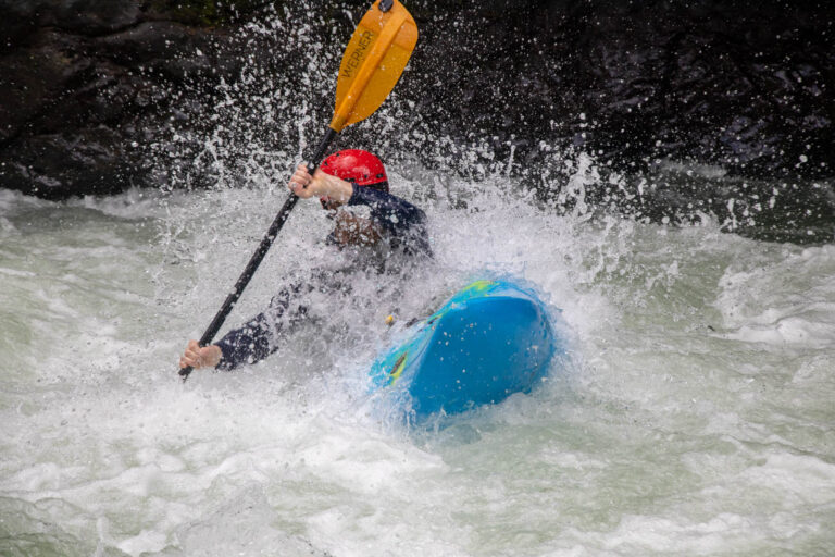 A kayaker in a red helmet punches a blue boat through an exploding wave, spray flying