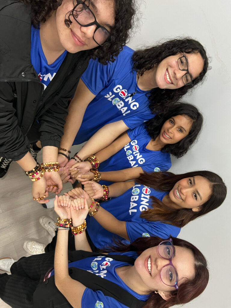 Five smiling teens in matching blue Going Global T-shirts hold out their wrists to show beaded bracelets
