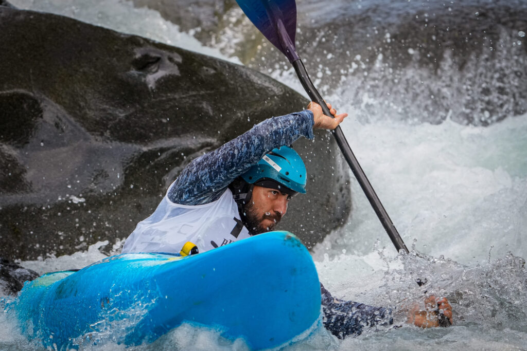 A bearded kayaker in a teal helmet powers a blue kayak through foaming whitewater beside a dark boulder