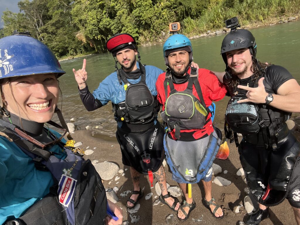 Four grinning kayakers in helmets and spray skirts take a selfie on a sandy riverbank beside a jungle river