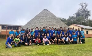 About fifty students in blue school shirts gather on a lawn in front of a tall cone-shaped thatched hut
