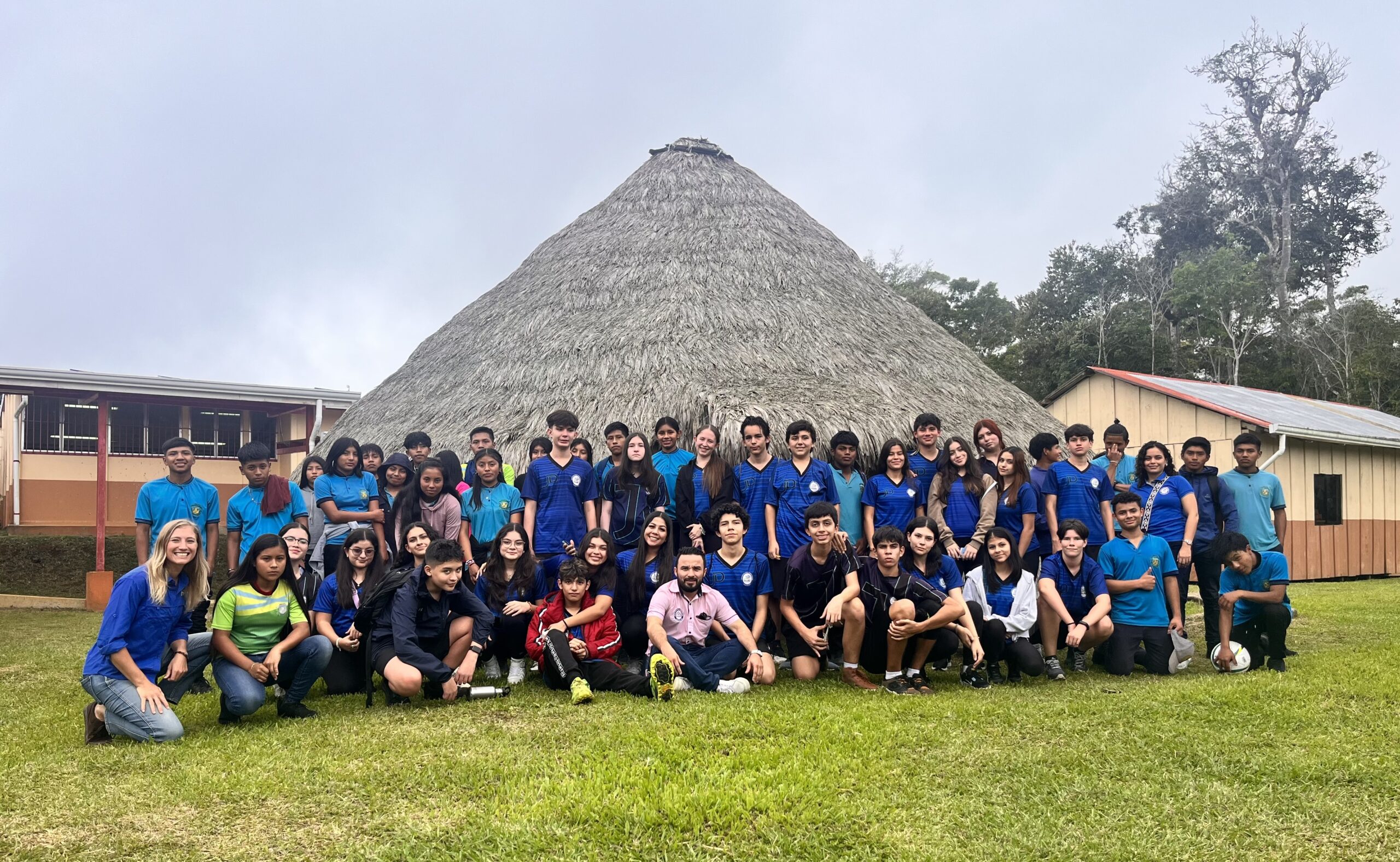About fifty students in blue school shirts gather on a lawn in front of a tall cone-shaped thatched hut