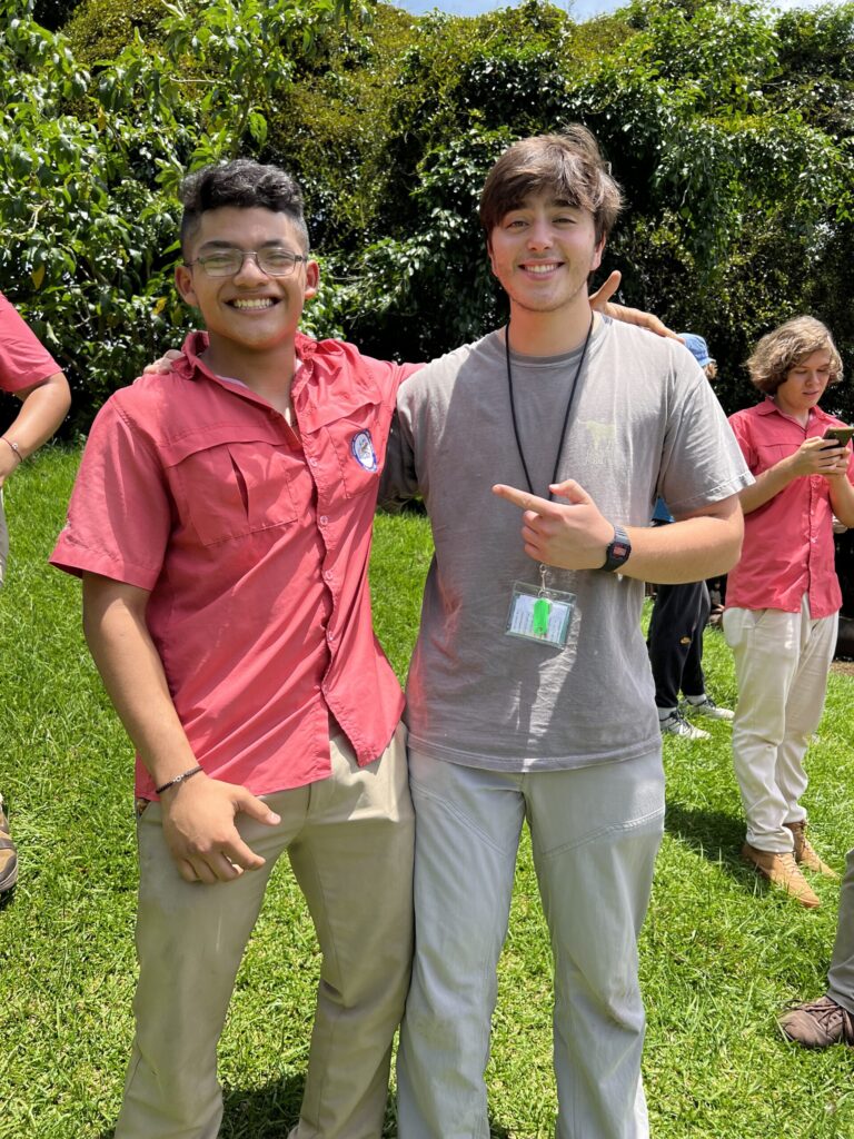 Two smiling teenage boys, one in a coral school shirt, pose arm in arm on a sunny lawn with classmates behind