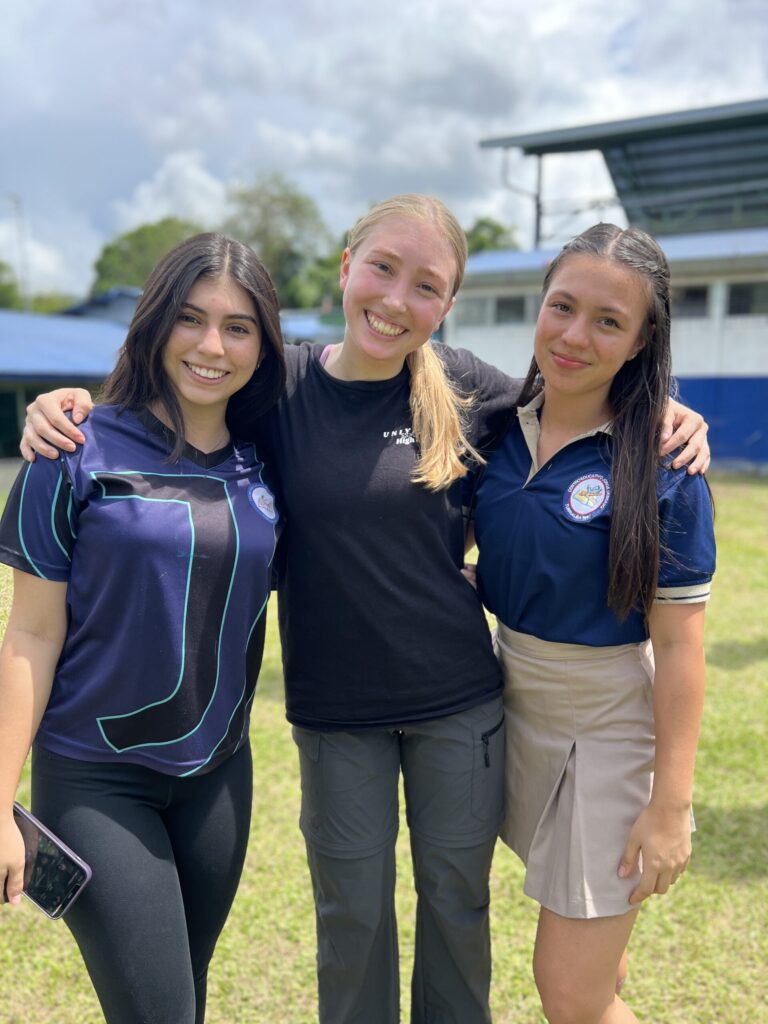 Three smiling teenage girls stand arm in arm on a school field, two of them in school uniforms