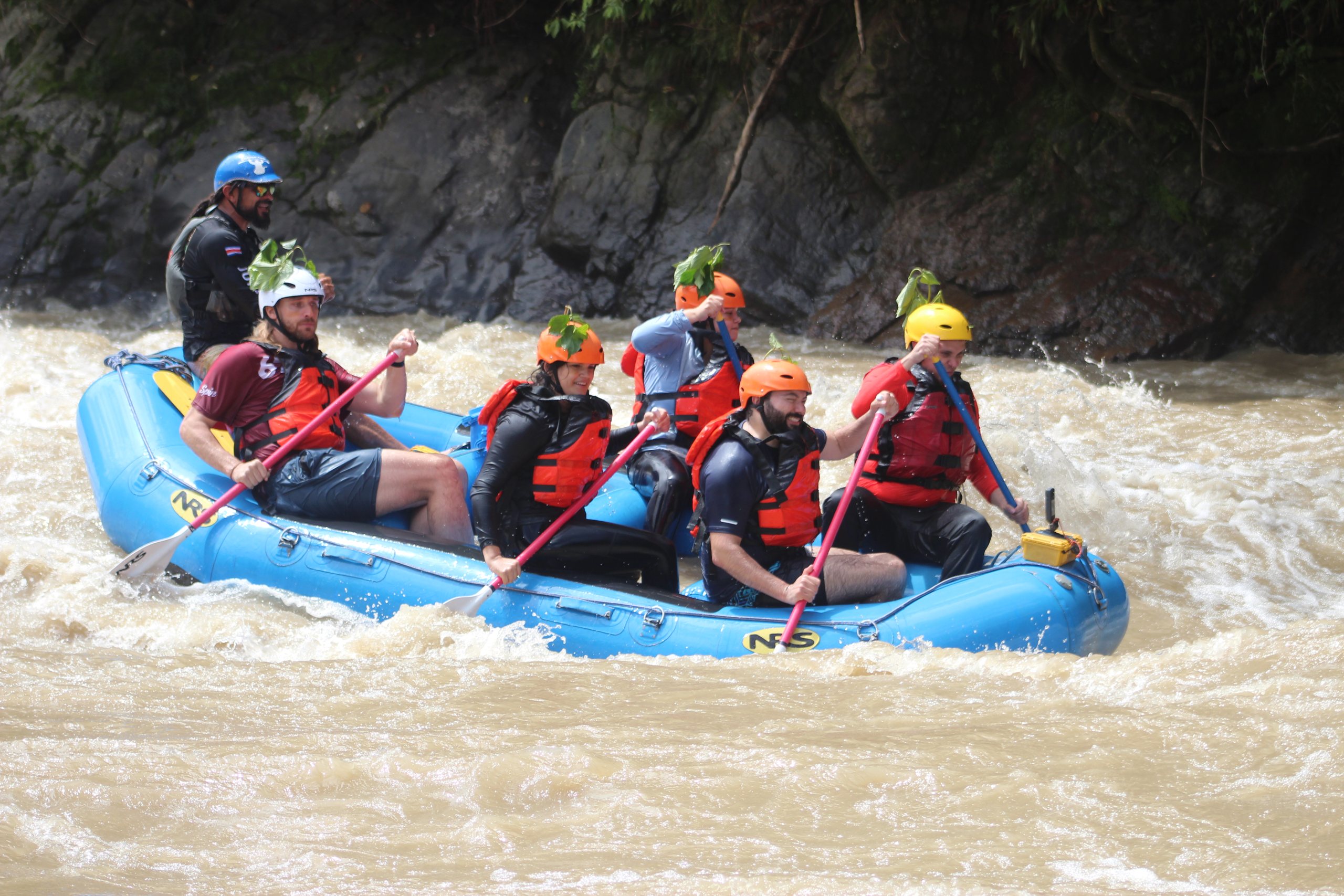 A blue raft of paddlers, some with leaves tucked into their helmets, works through brown whitewater on the Pacuare River