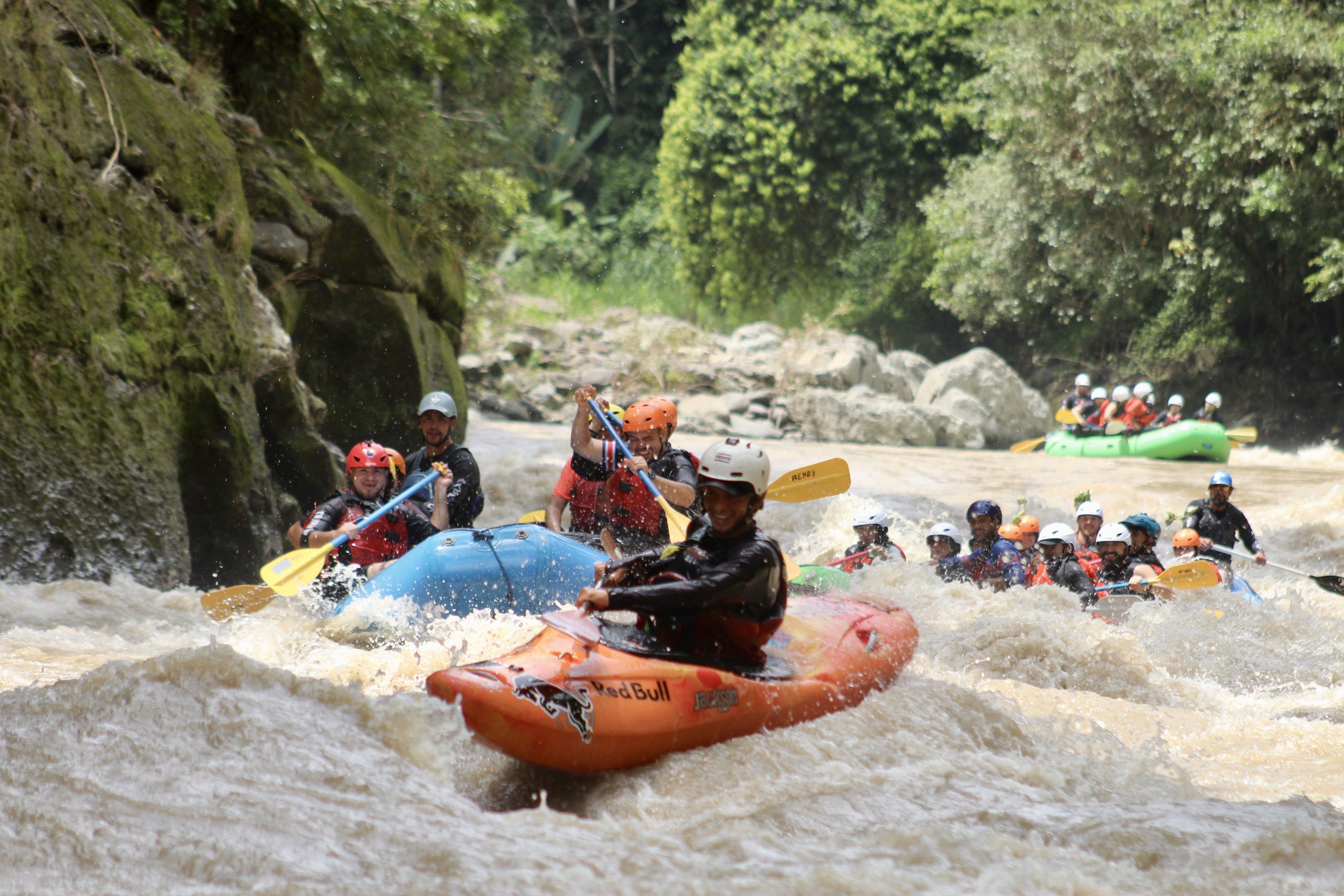 A kayaker in an orange boat leads rafts through churning whitewater between a mossy rock wall and the jungle on the Pacuare