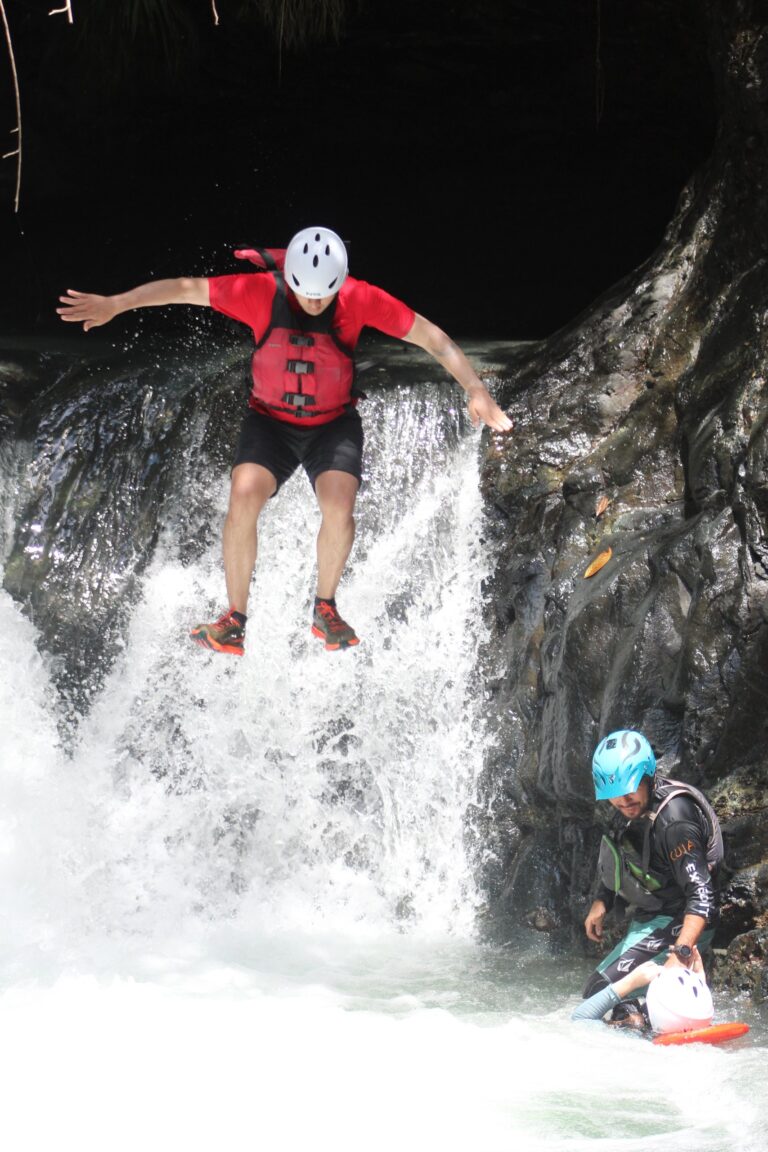 A rafter leaps from a waterfall into a clear pool as a guide in a blue helmet waits below with a rescue line
