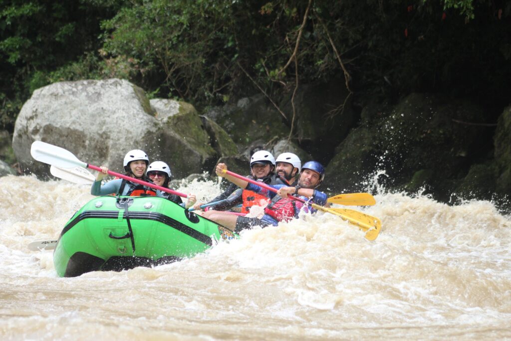 Five rafters in a green raft dig their paddles in as whitewater breaks over the bow beside mossy boulders