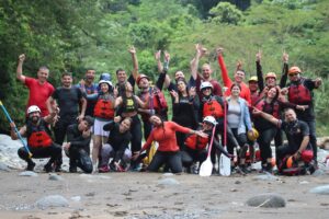 The rafting group celebrates on the riverbank with arms in the air, jungle-covered hills behind them